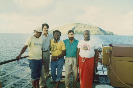 Colour photograph of five people standing on a ship at sea.