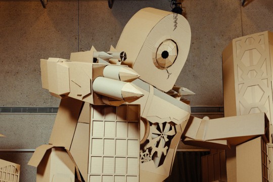 Giant cardboard robot sculpture holding on to cardboard buildings and standing on a cardboard vehicle