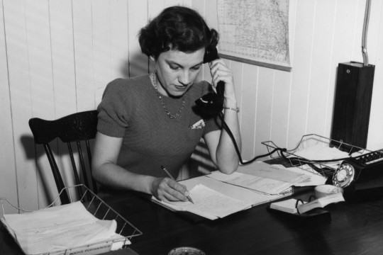 Black and white image of woman sitting at a desk writing while holding a telephone to her ear ca 1952