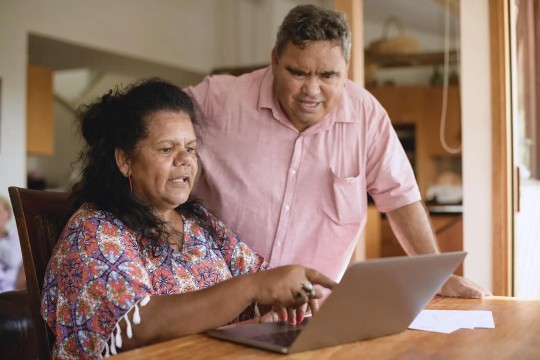 Woman is sitting and pointing at a laptop screen. A man stands behind her and watches.