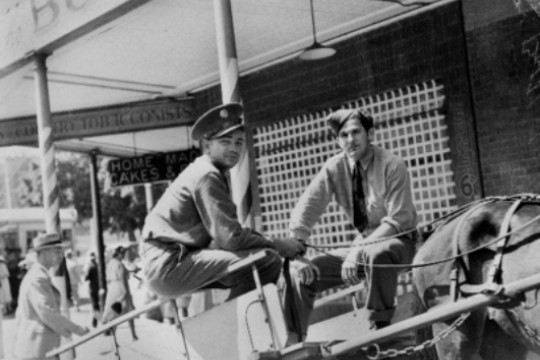 Two men sitting in a cart being pulled by a horse in a street with shops