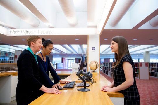 Visitor talking to two staff members at an information desk. One staff member is using a desktop computer.