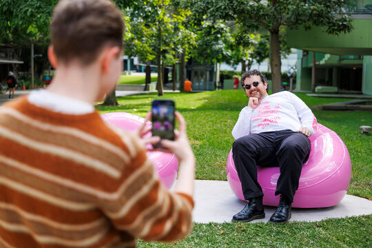 Man sitting on a Pink Chair outside, while someone takes a photograph. 