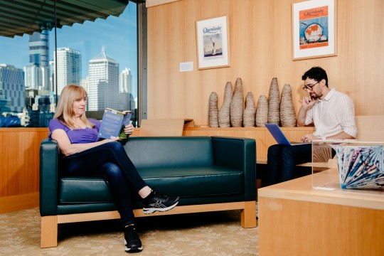 Two visitors in the John Oxley Library. One visitor reads a book on a couch while the other is using a laptop.