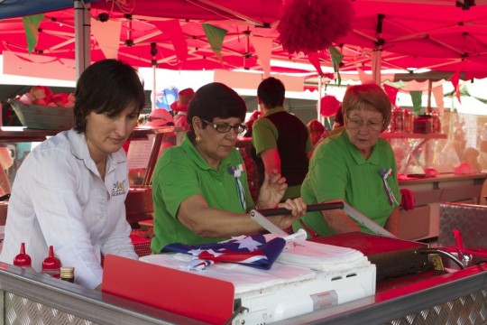 Three women are shown working at a delicatessen stall in a pavilion with a red roof Red white and green flags hang above