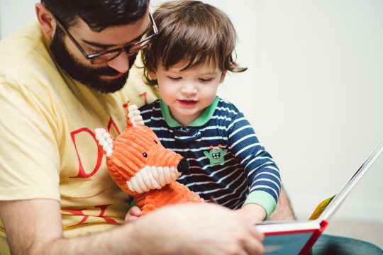 A man is reading from a book to a child he is holding. A stuffed toy fox is also on his lap.