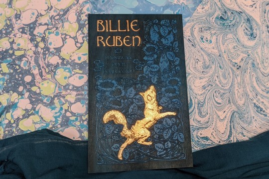 A laser-etched book cover dyed teal and embellished with gold leaf resting on a marbled-paper ground 