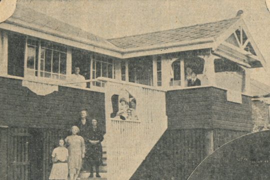 External view of Linga Longa two story house with several people standing on the stairs leading up to the second level
