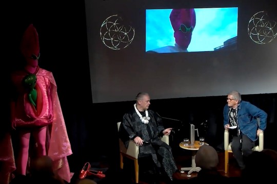 Two people sitting on stage with a projector screen and artwork.