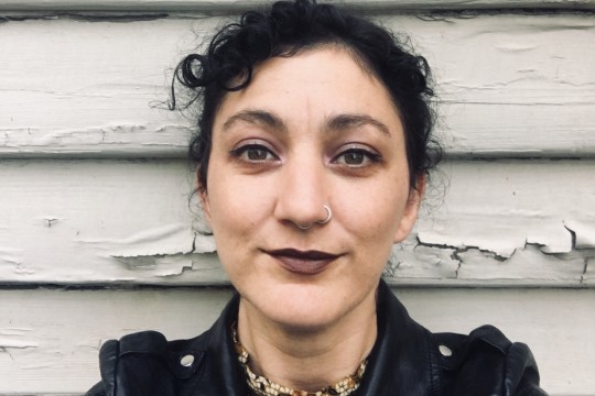 Mykaela Saunders takes a selfie in front of a white wall with peeling paint. She wears a black leather jacket and dark lipstick.