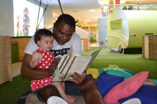 A man is reading to a toddler in a red dress who is sitting on his lap from a picture book They are sitting on brightly coloured cushions
