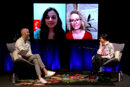 Two people sitting on stage with two people on large screens between them.