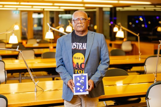 Lionel Fogarty at the 2023 Queensland Literary Awards. He stands in a study room with lamps. He is smiling and holding a copy of his book