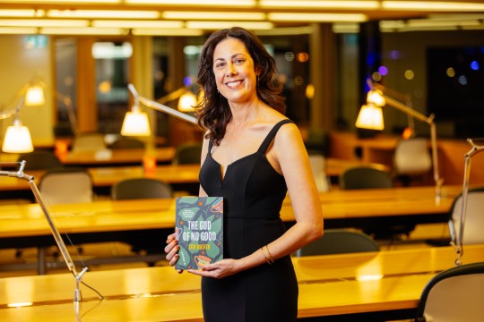 Sita Walker at the 2023 Queensland Literary Awards. She stands in a study room with lamps. She is smiling and holding a copy of her book.