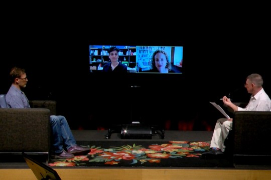 Two people sitting in chairs on a stage. Two people are seen on large screens between them.