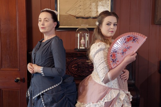 Two female actors wearing colonial dresses one in dark grey the other in pink and holding a fan