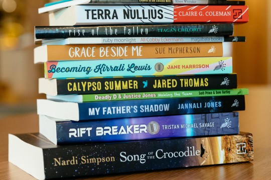 A collection of the black&write! published books in a pile. The books are sitting on a brown table. 