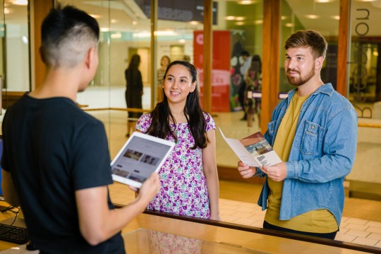 Two visitors talking to a State Library staff member at the reception desk One visitor is holding a brochure