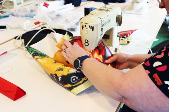 A women's arms are seen holding a brightly patterend piece of vinyl material, she is sitting in front of the showing machine and sewing the piece into a shape.