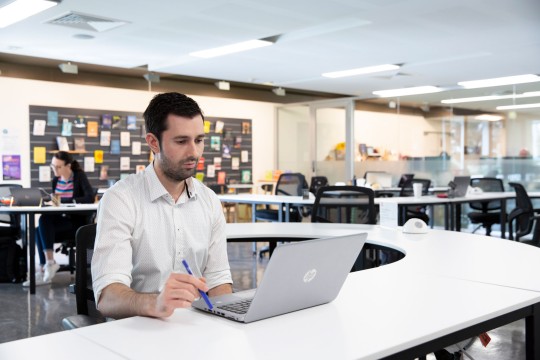 Man using laptop while holding a pen in a communal working space. 