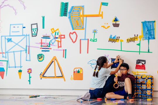 Two children using colourful marking tape to make pictues on a white wall.