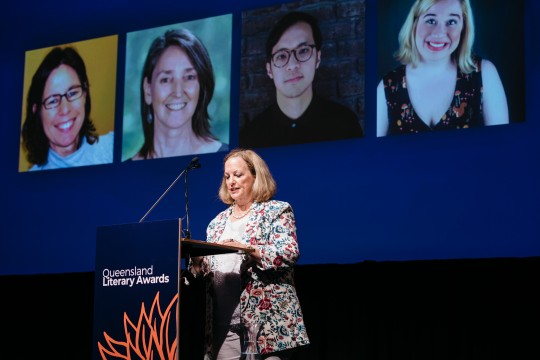 Woman standing and speaking at a podium with a projection behind her.