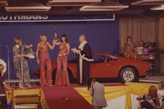 men and women on stage in front of a sports car accepting an award at a 1970s fashion showcase
