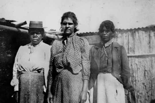 Three First Nations women standing outside a wooden shed