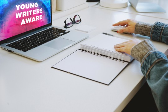 Hands on a mouse and a notepad. A laptop has Young Writers Award graphic on it.