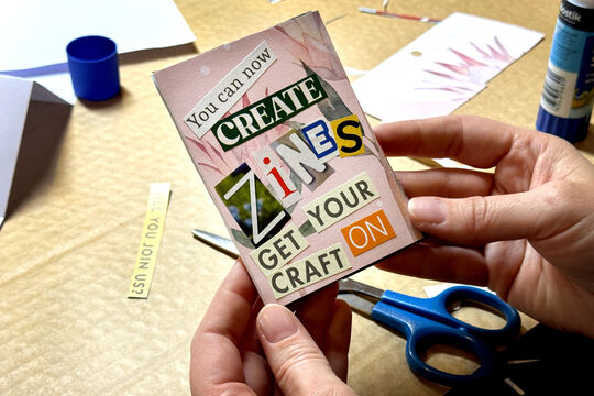 A person holds a handmade mini zine that says 'you can now create Zines' with cut out writing on a pink background