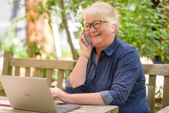 An elderly woman speaking on the phone while sitting at a table with a laptop.