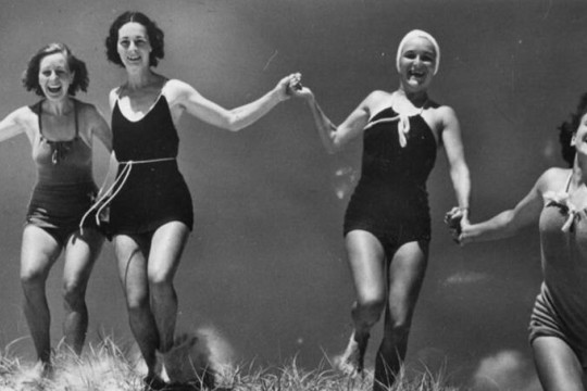 4 women running through grass in swimwear holding hands