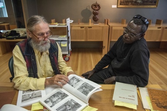 Two men with glasses in the Qld State Library looking through book with pictures