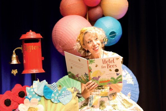 A picture of a woman in bright clothing on stage smiling at audience and holding a book hotel for bees
