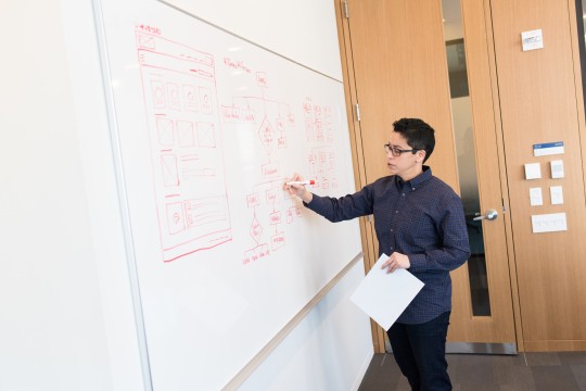 A person with short dark hair and glasses is writing on a whiteboard
