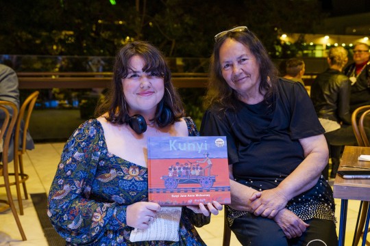 Two women sit beside each other at a night-time function. One holds up a copy of the book Kunyi by Kunyi June Anne McInerney