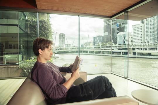 A young person sits in the Red Box at the library reading in an armchair. The river is behind him.