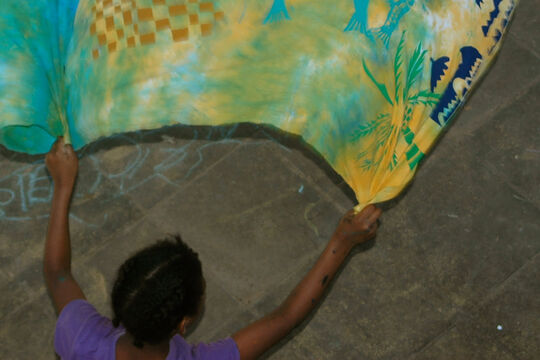 A child holding up a large, colourful fabric decorated with fish, palm trees, woven patterns, and abstract shapes in shades of blue, green, yellow, and orange.