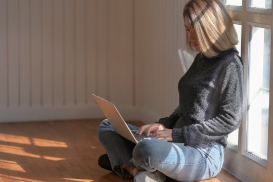 Woman in grey sweater is sitting on the floor and looking at a laptop