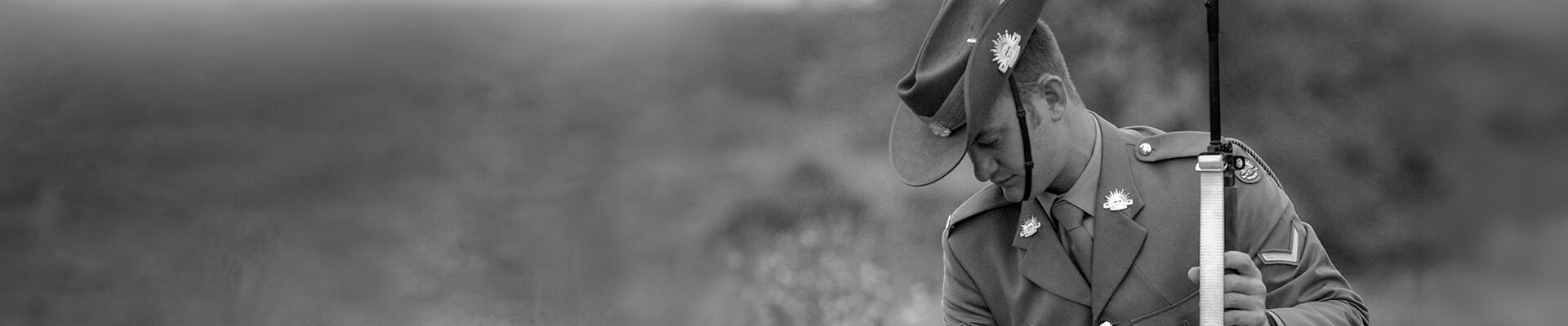 Black and white photo of a solider kneeling tp pick a poppy from a grassy field Poppy flowers have been coloured red and are the only colour in the photo