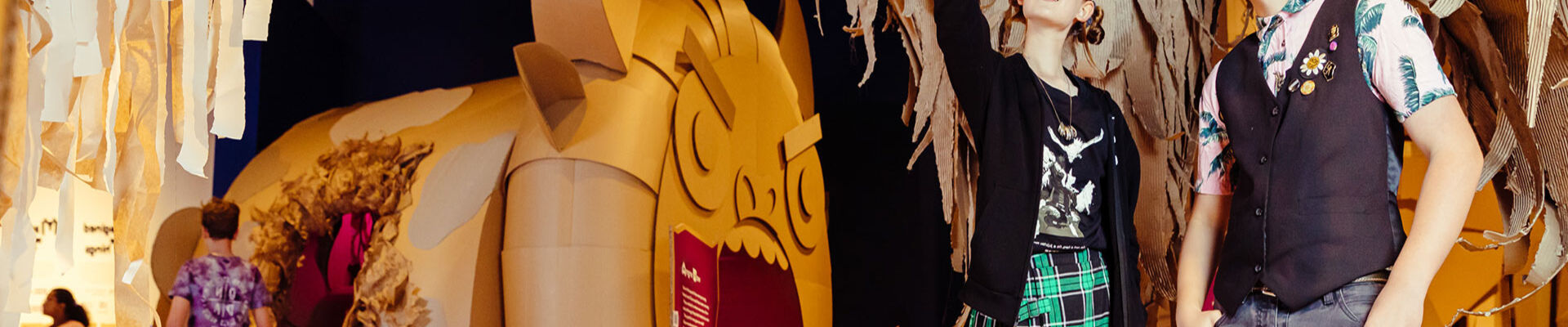 Two teens standing and pointing in front of large cardboard sculptures at State Librarys The Great Grand Rumpus exhibition