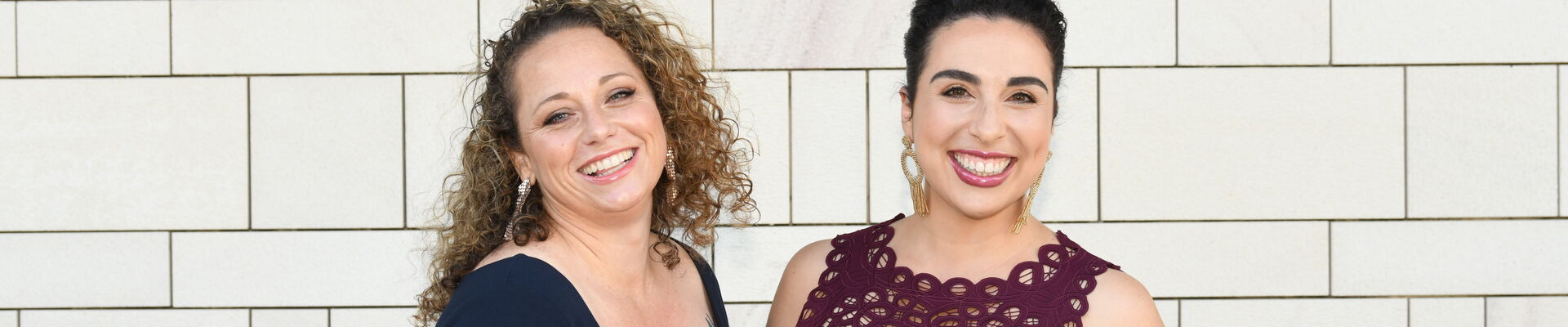 Like A Photon Creative Kristen Souvlis and Nadine Bates from Like A Photon Creative standing together in front of a white brick wall and smiling at the camera