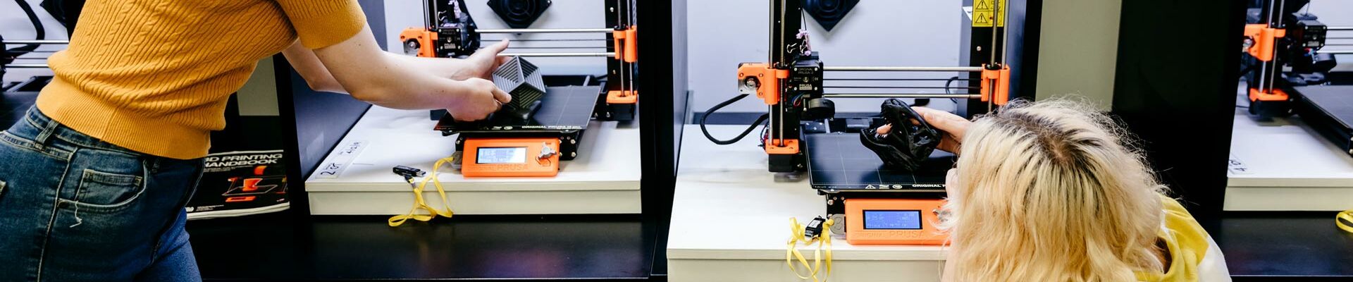 Two people using 3D printers One person is crouched down examining a creation and the other is standing