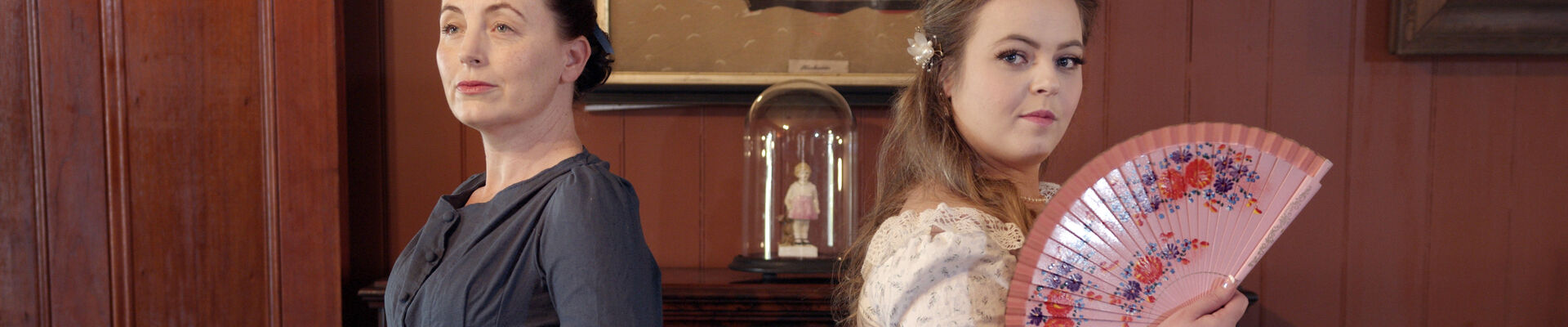 Two female actors wearing colonial dresses one in dark grey the other in pink and holding a fan