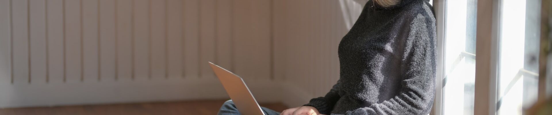 Woman in grey sweater is sitting on the floor and looking at a laptop
