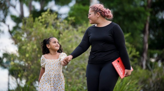 Mother and daughter walking outside holding hands. Mother is holding a book titled Koalas Like To ... written by Written by Shae Millward.