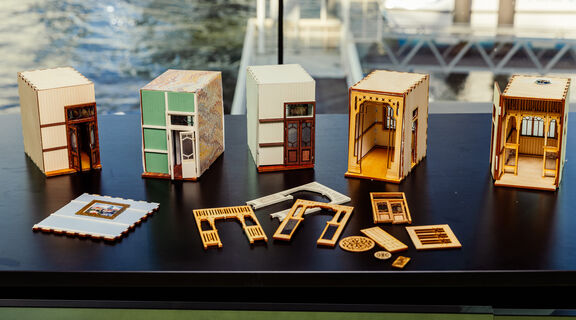 A row of detailed miniature house models made from wood and paper sits on a black table with various decorative parts laid out in front of them. The background shows a waterfront scene through a large window.