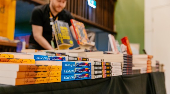 A person stands behind a table with piles of books on it at a pop-up books shop.