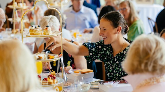 Person using tongs to pick up a small cake from a high tea tray.