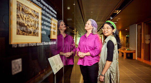 Two women looking at artworks hanging on a wall behind glass.